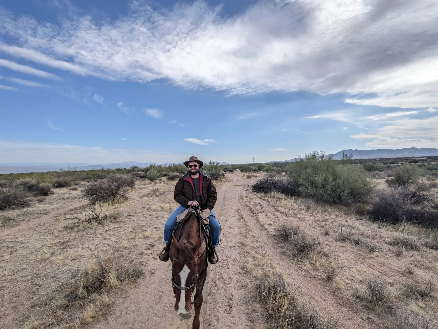 On horseback on a desert trail, sunglasses on, looking into the camera.
